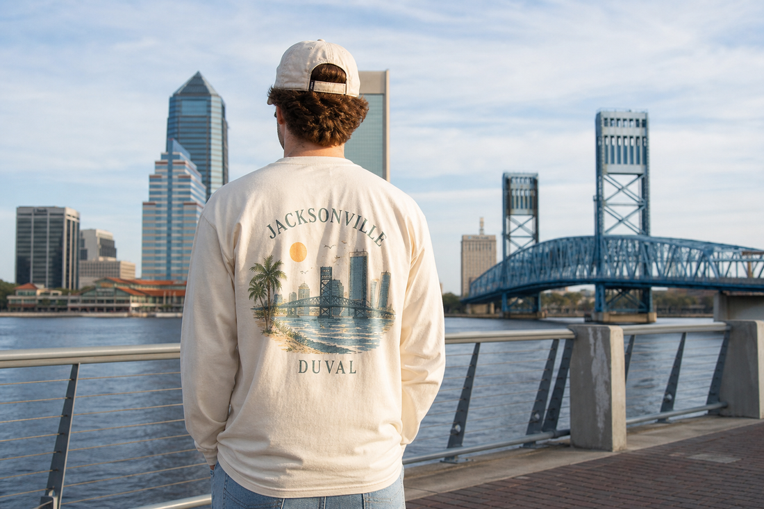 Person wearing a Jacksonville-inspired long sleeve streetwear shirt near the river with a bridge and skyline in the background.